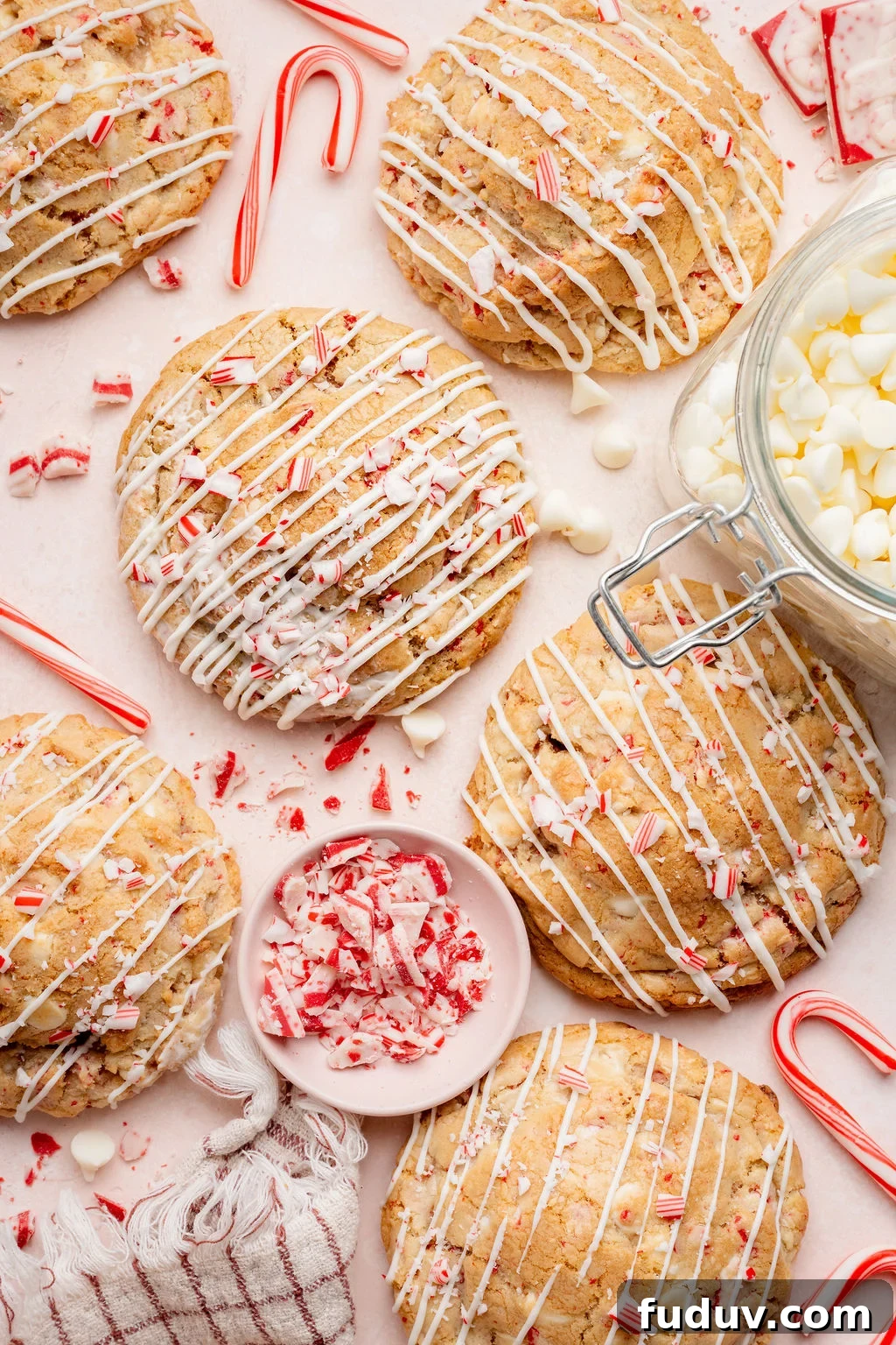 White Chocolate Peppermint Cookies Recipe for the Holidays 2 Overhead view of large white chocolate peppermint cookies drizzled with white chocolate and topped with crushed candy canes, surrounded by candy cane pieces, white chocolate chips, and a small bowl of peppermint bark on a light pink surface.
