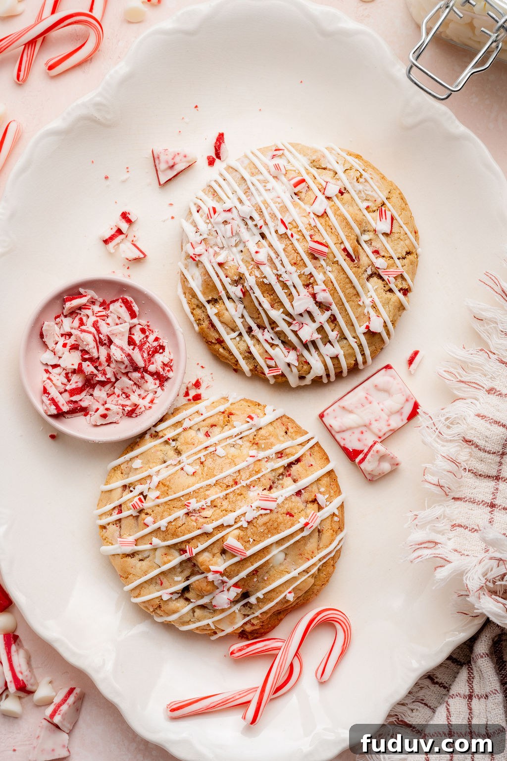 White Chocolate Peppermint Cookies Recipe for the Holidays 13 Overhead view of two large white chocolate peppermint cookies on a scalloped white platter, each drizzled with white chocolate and topped with crushed candy canes. A small pink bowl filled with chopped peppermint bark sits beside the cookies, with scattered candy cane pieces and a striped kitchen towel adding festive detail around the tray.