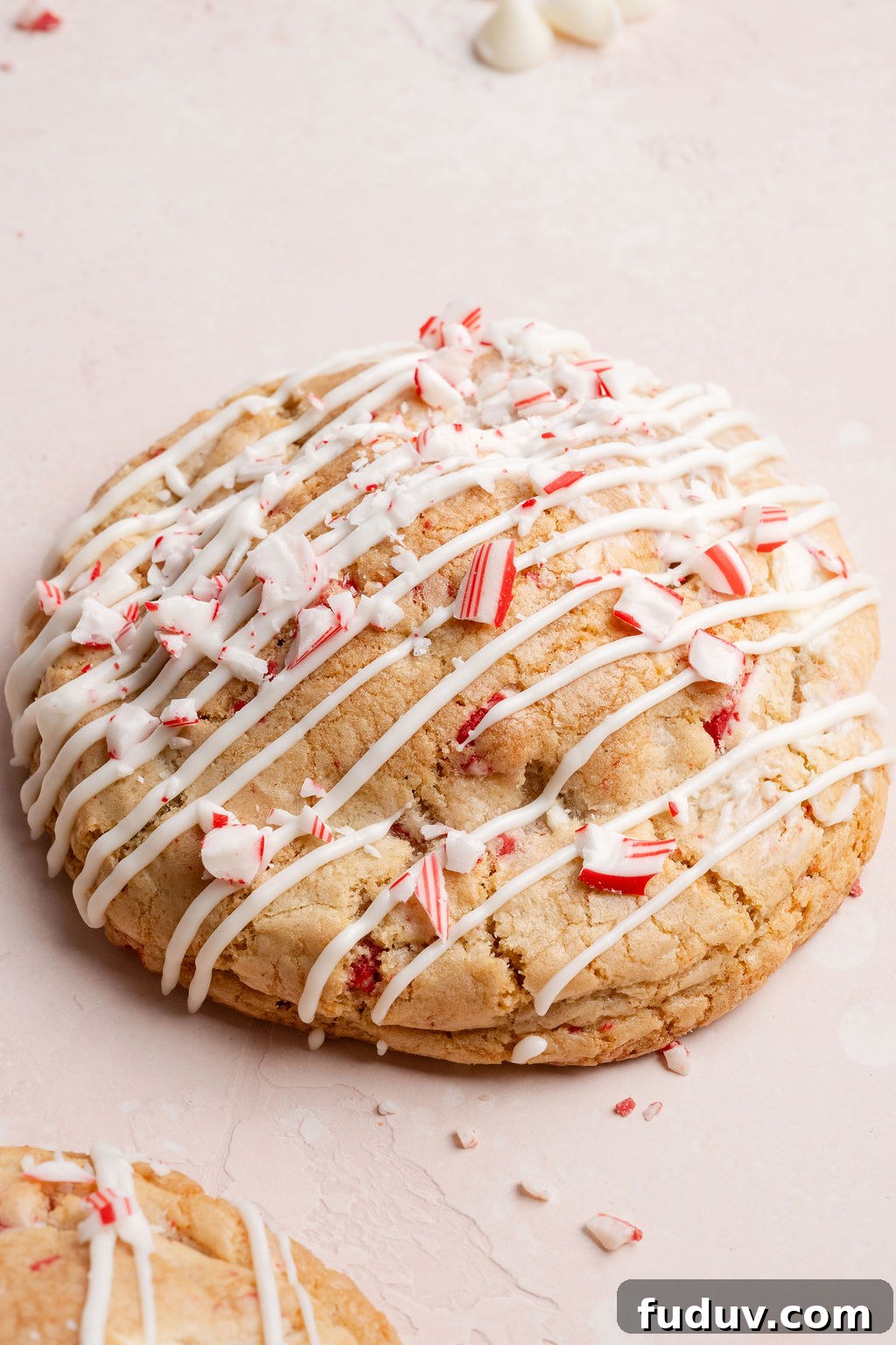 White Chocolate Peppermint Cookies Recipe for the Holidays 6 Close-up of a single white chocolate peppermint cookie topped with white chocolate drizzle and crushed candy canes on a light pink surface.
