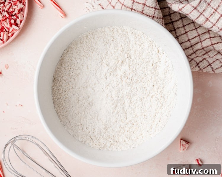 Large white mixing bowl filled with whisked dry ingredients for cookie dough, surrounded by peppermint pieces, a whisk, and a kitchen towel on a pink surface.