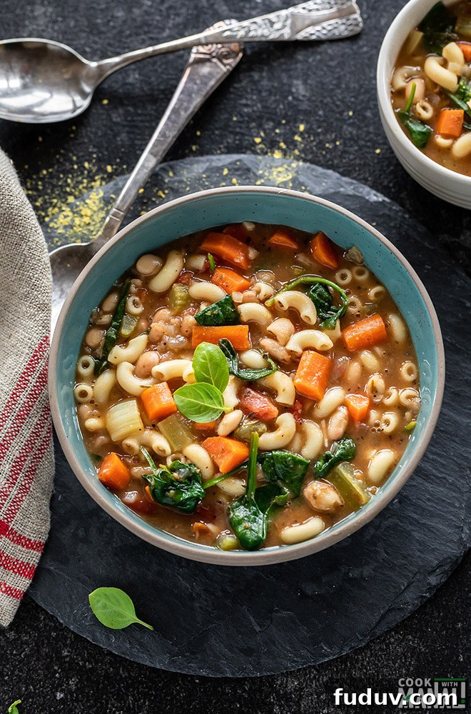 bowl of minestrone soup with two spoons in the background and a kitchen towel on the side