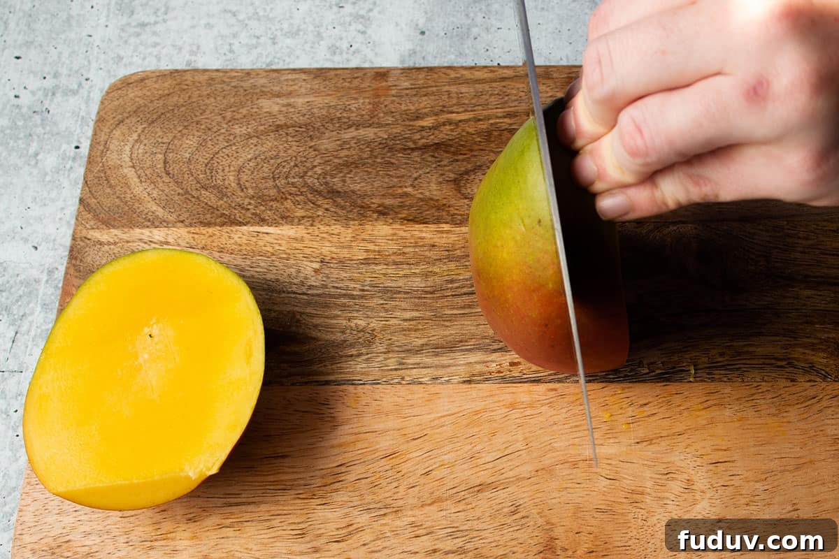 A knife cutting the second side of a mango with the fist side removed.