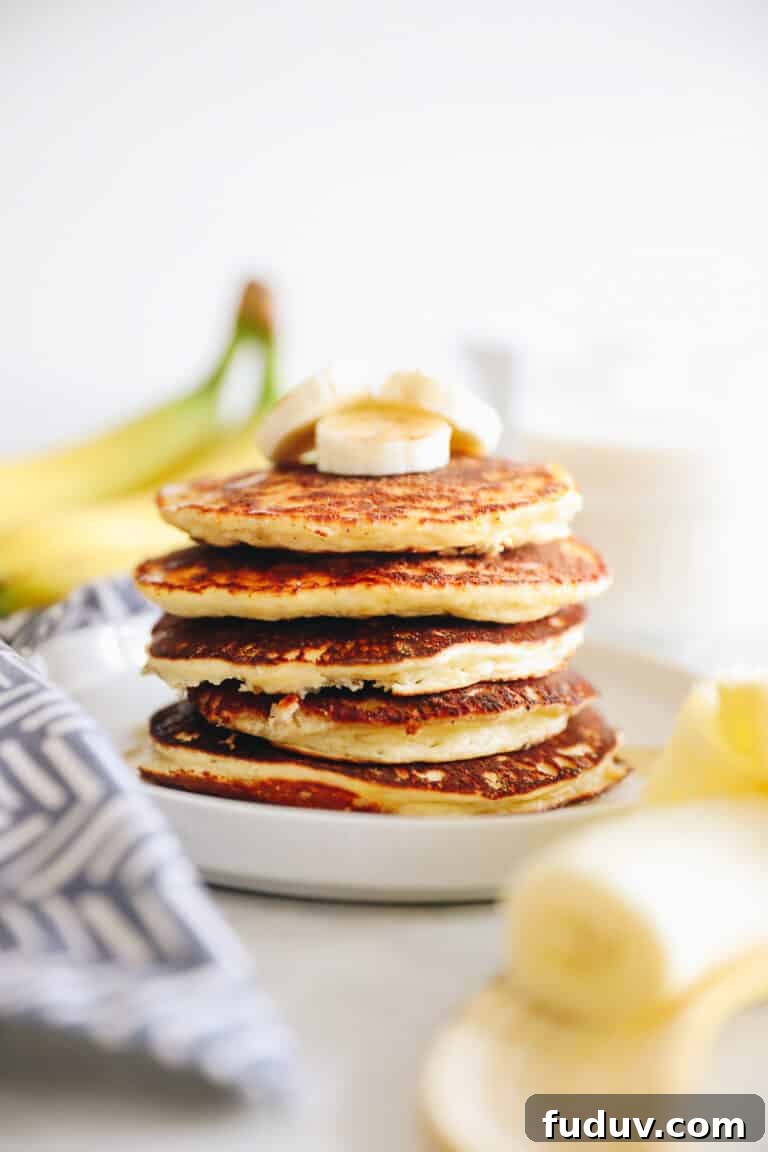 Banana protein pancakes stacked on a white plate. Bananas in foreground and background of the image.