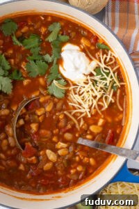 30+ Delicious Recipes to Transform Leftover Turkey 7 A pot filled with hearty chili, featuring beans, turkey, and vegetables in a rich orange-red sauce. Topped with a dollop of sour cream, shredded cheese, and cilantro. A ladle rests in the pot, and a napkin and cornbread are visible nearby.