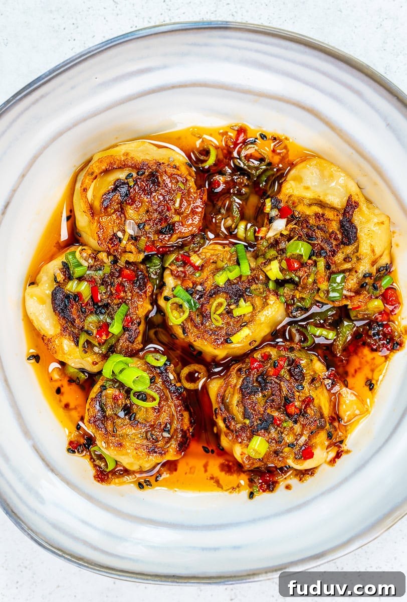 Crispy Homemade Dumpling Rolls: Step-by-Step Recipe 2 Six pan-fried dumplings topped with chopped green onions and red chilies sit in a white bowl, surrounded by a spicy chili oil sauce with visible pepper flakes and herbs.