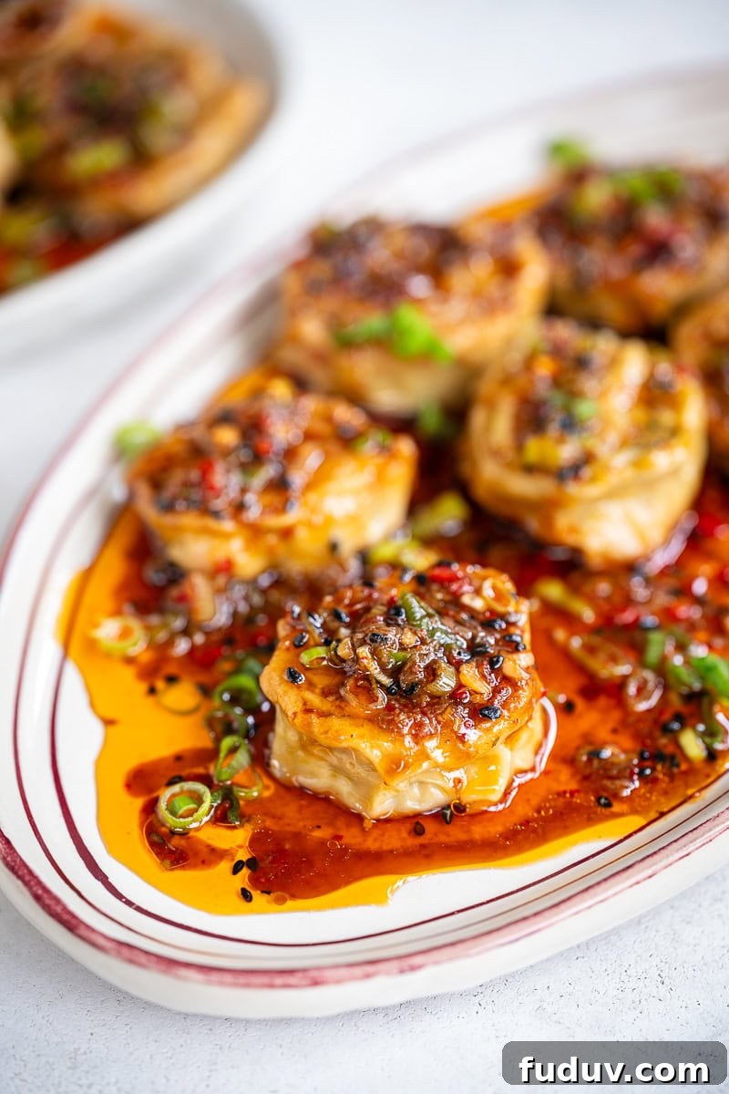 Crispy Homemade Dumpling Rolls: Step-by-Step Recipe 3 A plate of dumpling rolls topped with a savory sauce, garnished with chopped green onions and sesame seeds, sits on a white dish with red trim. The sauce is glossy and vibrant, pooling around the tofu pieces.