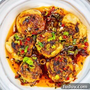 Crispy Homemade Dumpling Rolls: Step-by-Step Recipe 10 Six pan-fried dumplings topped with chopped green onions and red chilies sit in a white bowl, surrounded by a spicy chili oil sauce with visible pepper flakes and herbs.