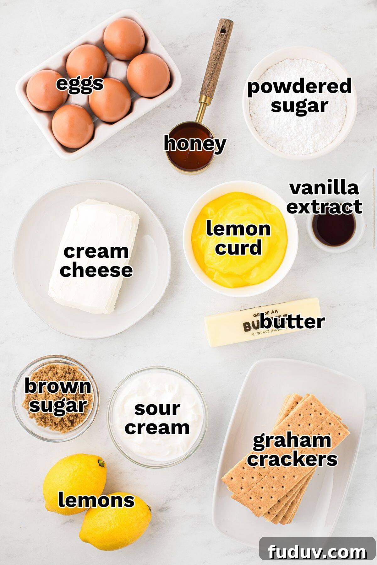 Various labeled ingredients for a lemon cheesecake recipe, including eggs, honey, powdered sugar, cream cheese, lemon curd, vanilla extract, butter, brown sugar, sour cream, graham crackers, and fresh lemons.