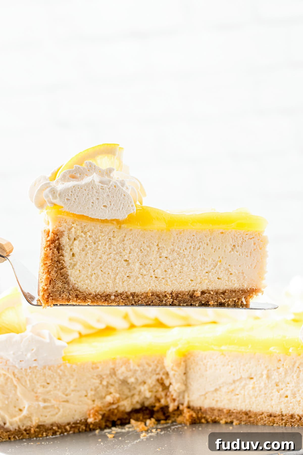 A slice of Lemon Cheesecake with a graham cracker crust, topped with lemon curd, whipped cream, and a lemon slice, held above the rest—this Lemon Dessert is sure to brighten any occasion.