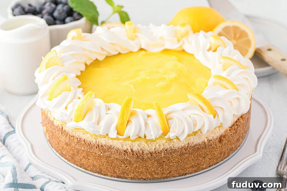 A Lemon Cheesecake topped with tangy lemon curd, whipped cream, and lemon slices, sits on a white plate with fresh lemon, knife, and blueberries in the background.