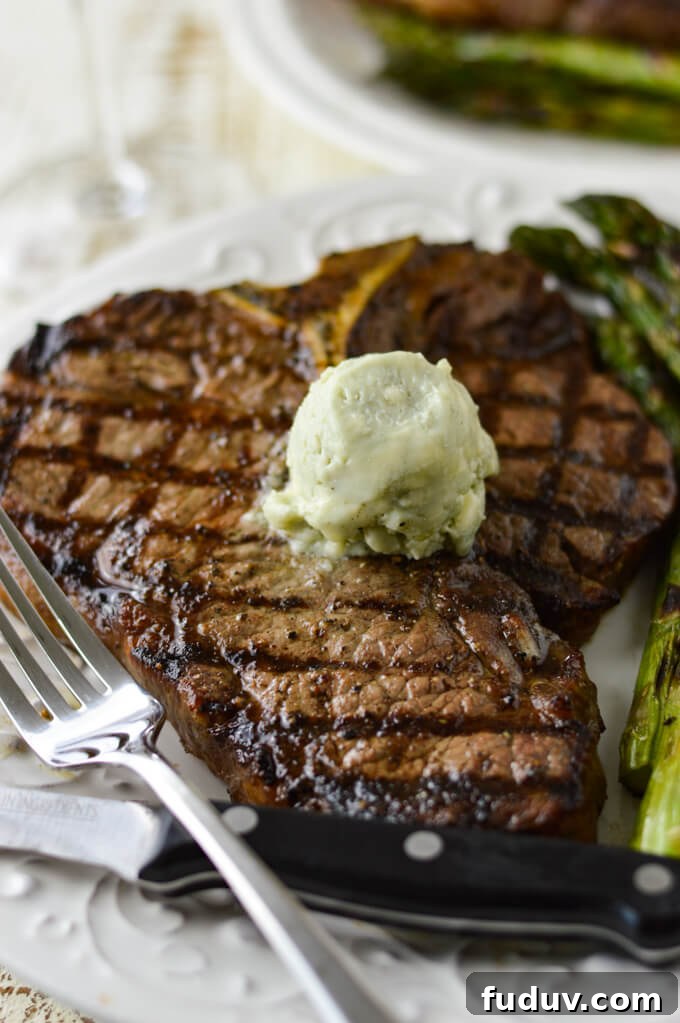 A grilled t-bone steak on a white plate with a round of blue-cheese butter melting on top. A fork and a steak knife sit in front and grilled asparagus is on the plate next to the steak.