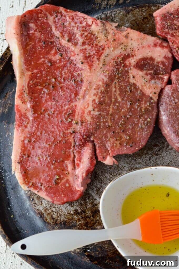 A raw t-bone steak showing the strip steak side and the tenderloin side with arrows sits on a pan with a small bowl of olive oil and an orange basting brush.