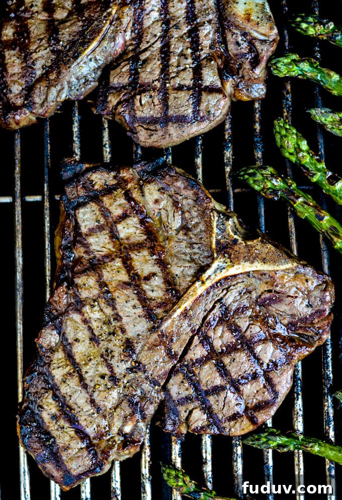 Two cooked t-bone steaks are on the grill with crisscross grill marks on them. Asparagus sits next to them on the grill.