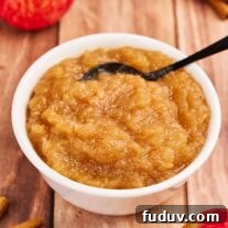 Homemade Cinnamon Applesauce Recipe: Warm Spice & Comfort 7 A round white bowl filled with cinnamon applesauce, with a black serving spoon, next to cinnamon sticks and whole, fresh apples on a rustic wooden tabletop.