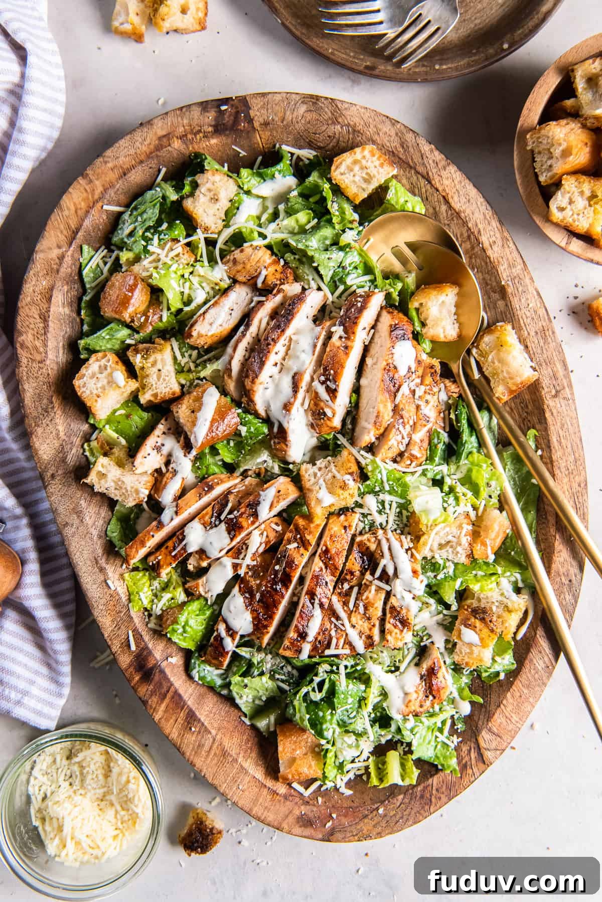 A top down shot of two salad spoons resting in a wood bowl filled with grilled chicken Caesar salad.