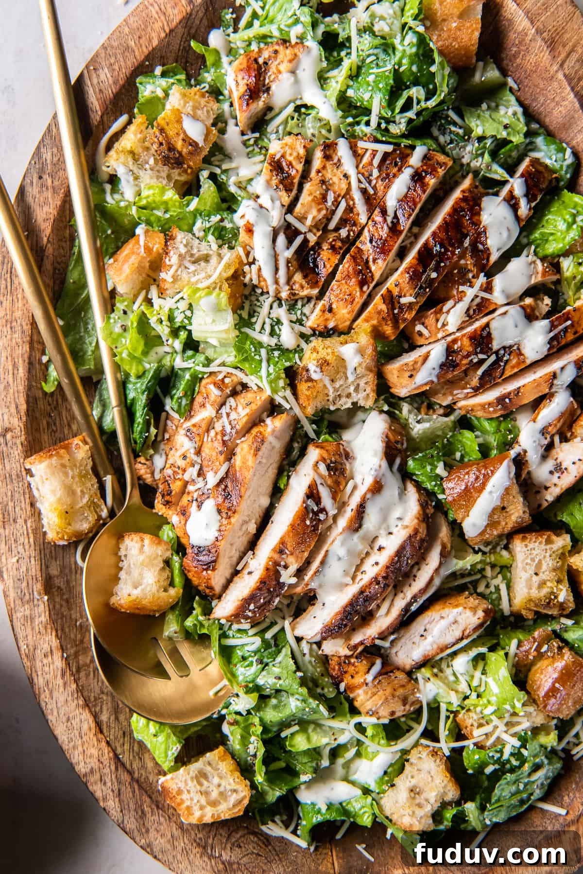 A top down close up shot of two salad spoons resting in a wood bowl filled with grilled chicken Caesar salad.