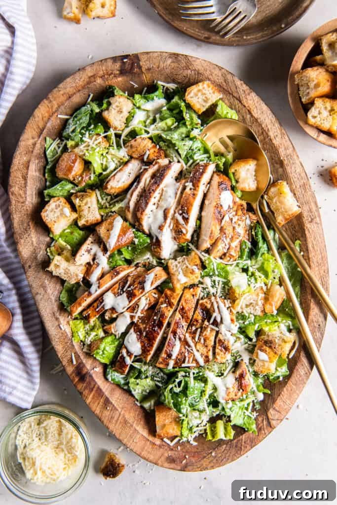 A top down shot of two salad spoons resting in a wood bowl filled with grilled chicken Caesar salad.