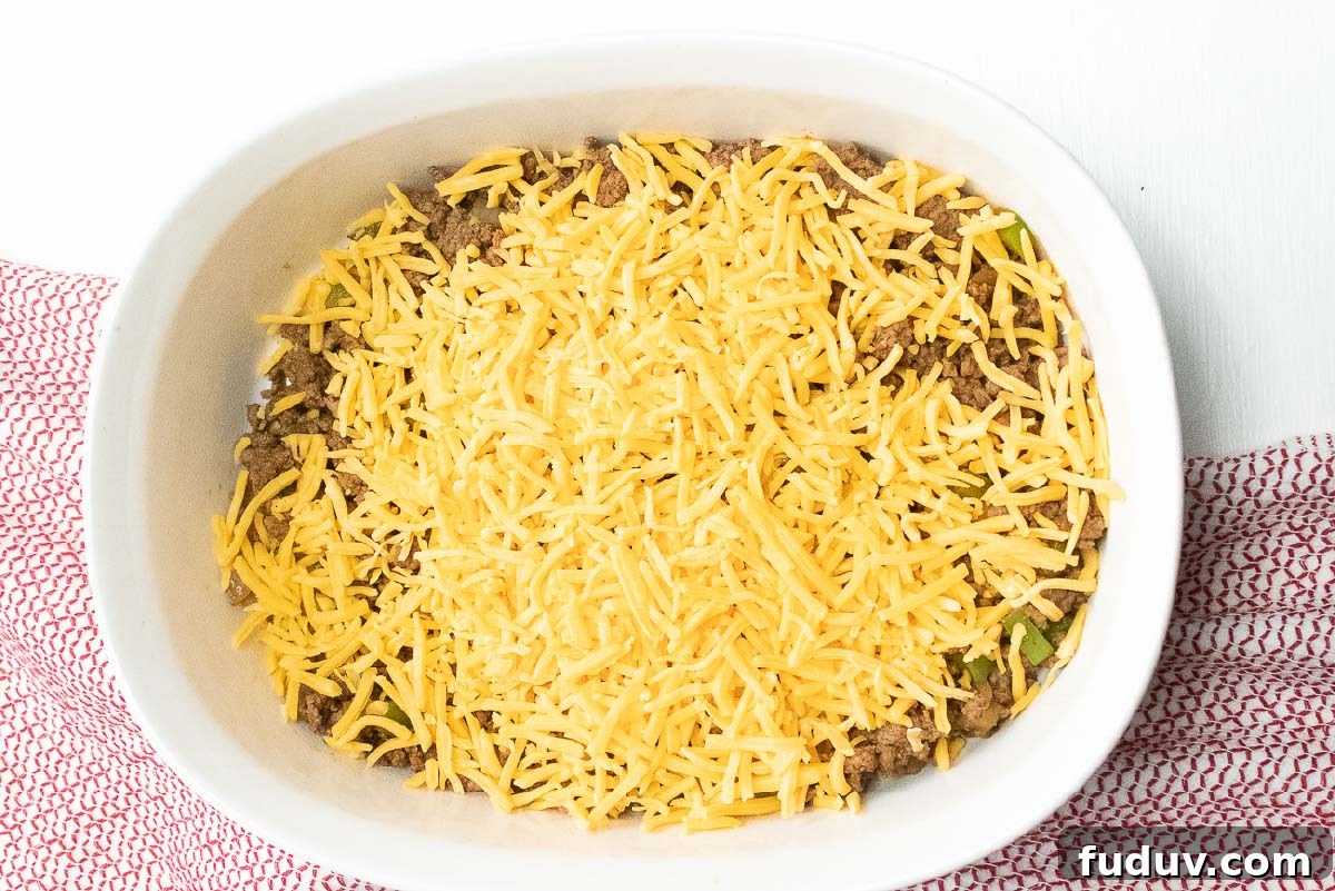 Cooked ground beef, diced onions and bell peppers in a white casserole dish, topped with shredded cheddar cheese.