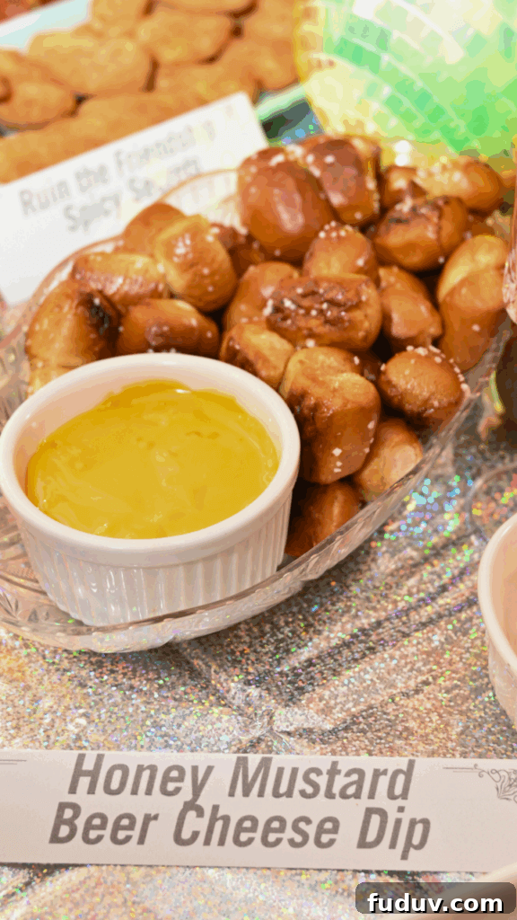 A close up of a small white bowl of honey mustard beer cheese dip surrounded by soft pretzel nuggets in a clear dish over an iridescent table cloth