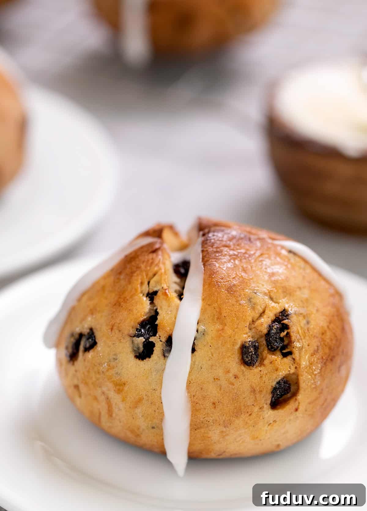 Hot cross bun with frosting cross on small white plate with butter in background