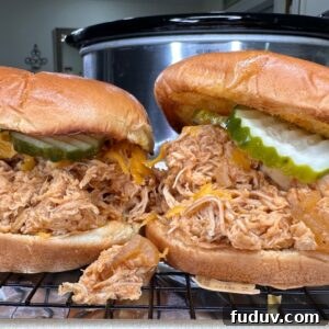 Two shredded chicken sandwiches on buns, topped with pickles and melted cheese, sit on a wire rack in front of a slow cooker. Some chicken filling spills out from the sandwiches.