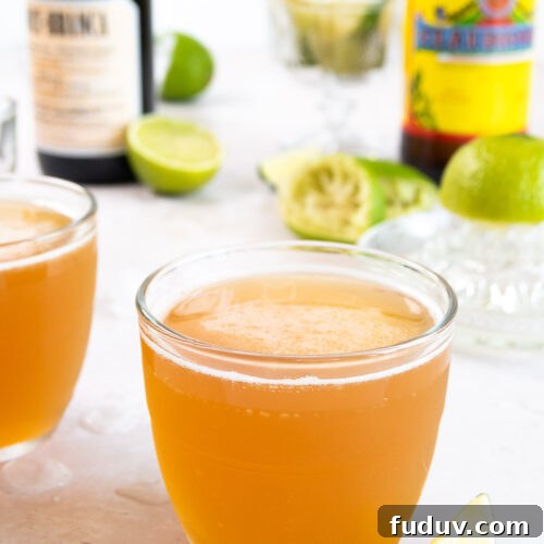 close up image of a Macunaíma cocktail in a small glass on a white surface in front of bottles and limes