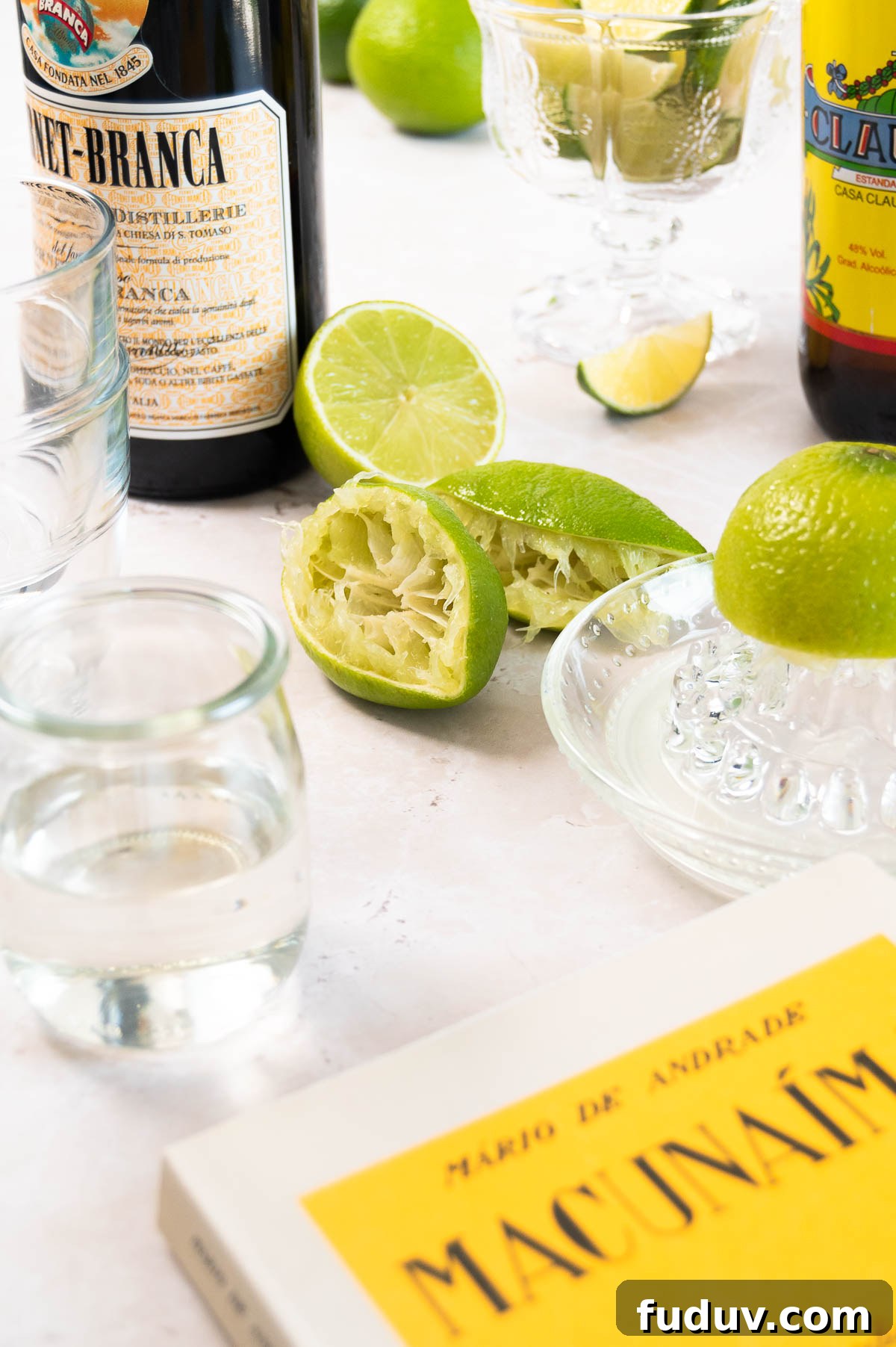 juiced limes with a glass citrus juicer, some liquor bottles and the edge of the Macunaíma book on a white surface
