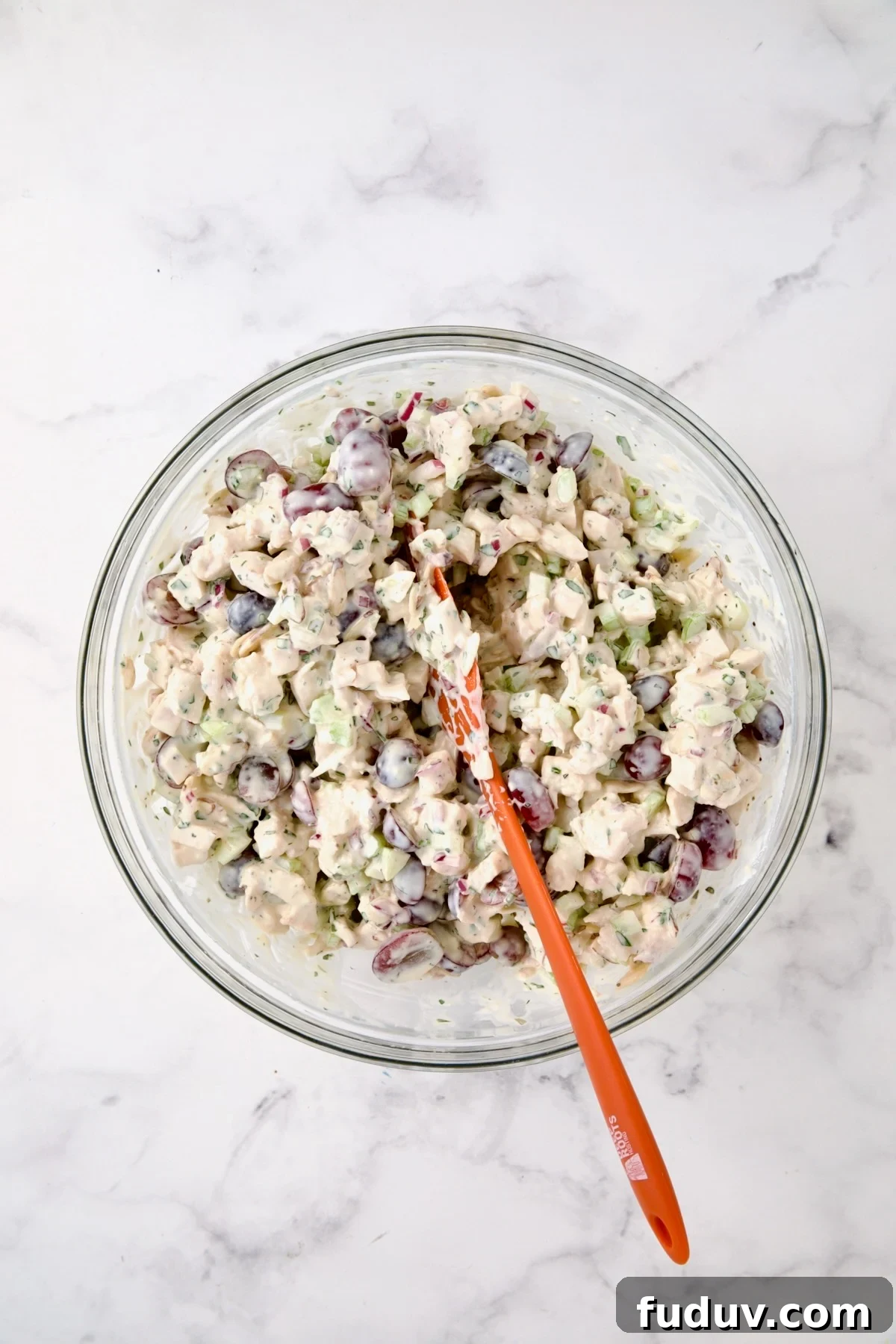 Chicken salad in glass bowl being stirred with orange spatula.